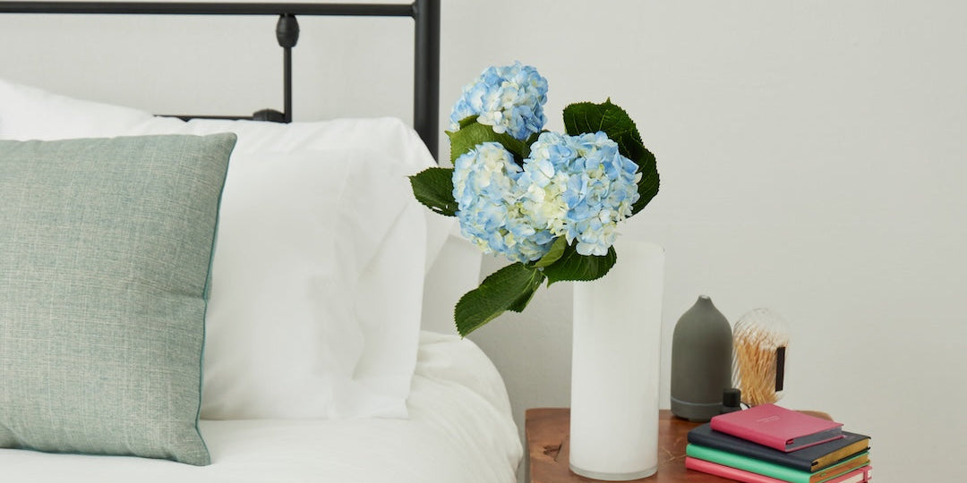 A styled bedroom corner showing a white bed with a black metal headboard, a white nightstand with a live-edge wood finish, blue hydrangeas in a white vase, stacked books, a small gray vase, and beige slippers on a woven rug.