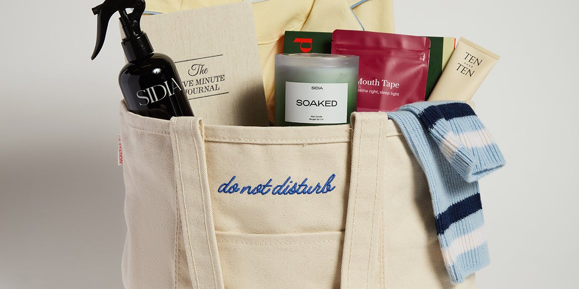 Beige tote bag with various items including pajamas, bath products, and boxes on a light gray background