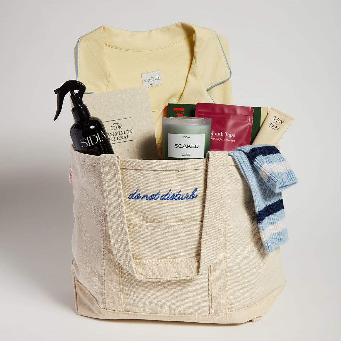 Beige tote bag with various items including pajamas, bath products, and boxes on a light gray background