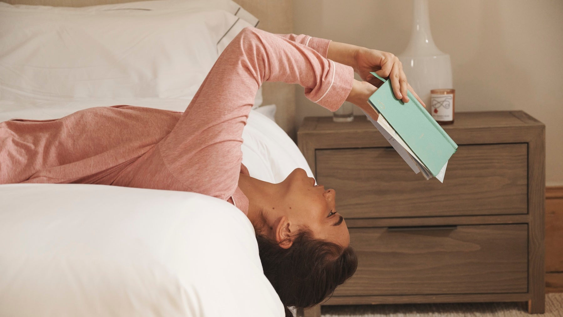 Woman lying on a bed holding a green book next to a wooden nightstand.