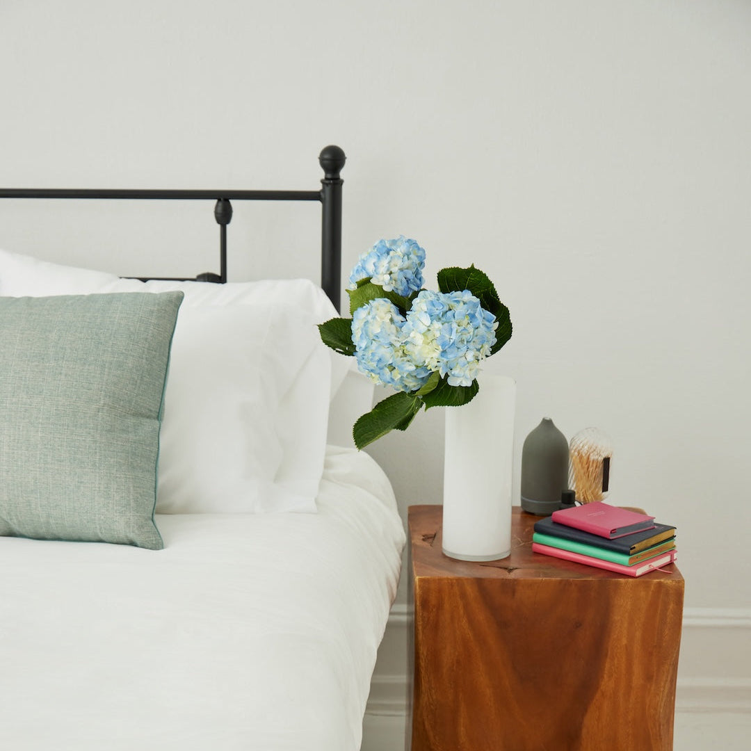 A styled bedroom corner showing a white bed with a black metal headboard, a white nightstand with a live-edge wood finish, blue hydrangeas in a white vase, stacked books, a small gray vase, and beige slippers on a woven rug.