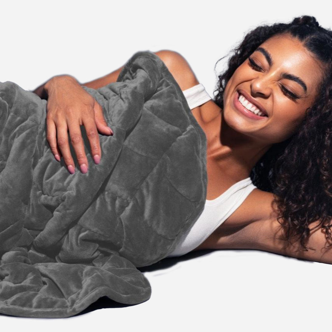 Woman lying on a white surface under a gray weighted blanket, smiling.