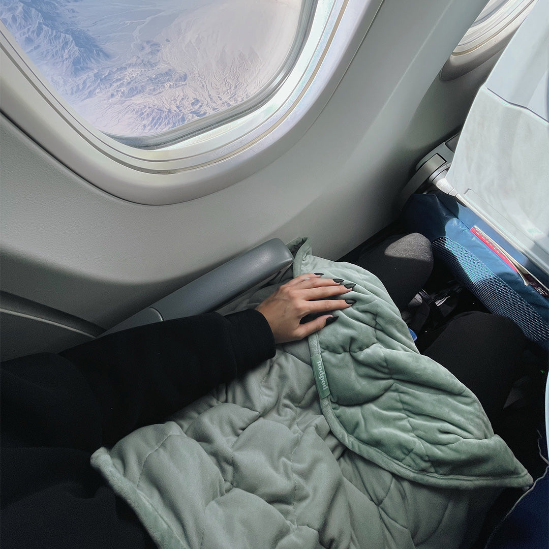 Hand touching a sage green weighted blanket on an airplane seat with a window view.