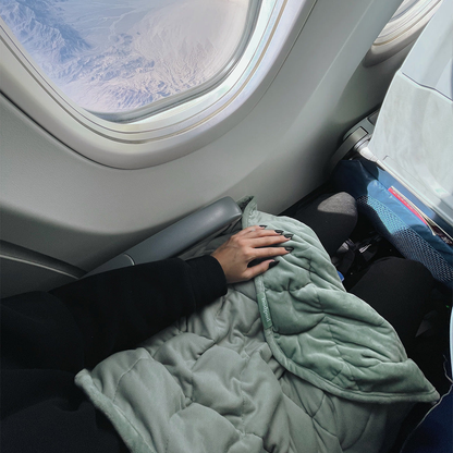 Hand touching a sage green weighted blanket on an airplane seat with a window view.
