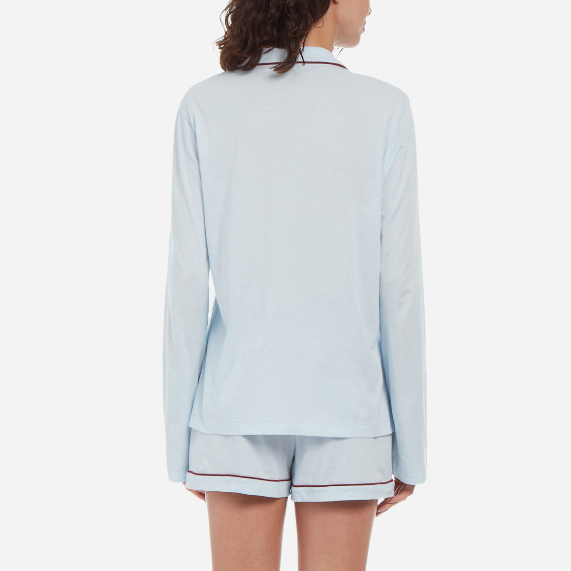  Back facing female model wearing long sleeve light blue pajama top with deep red piping with matching shorts.