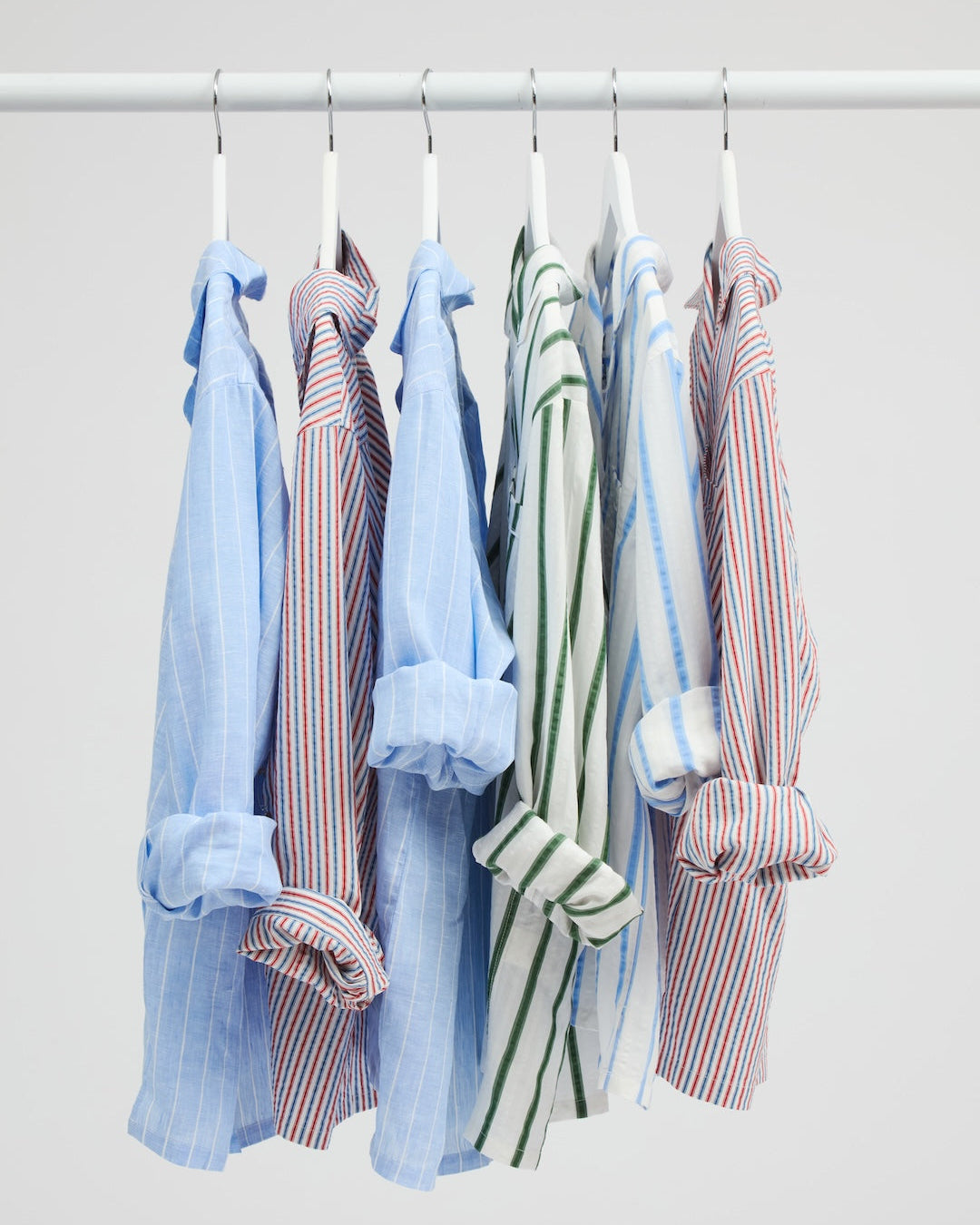 Six long-sleeve button-up shirts in various colors and stripe patterns hang on white hangers from a white horizontal rack against a light gray background.