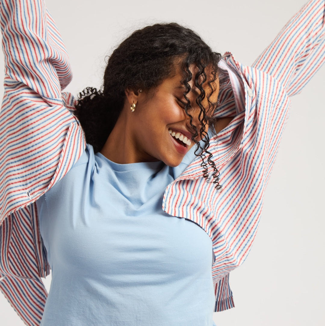 A woman wearing a light blue cropped t-shirt and a red, white, and blue striped button-up shirt with her arms raised, set against a plain light gray background.