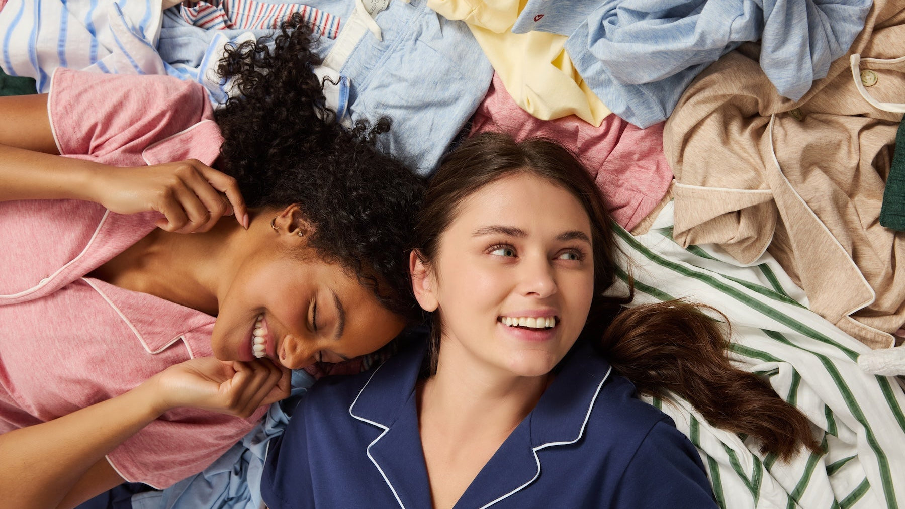 Two women lying on a bed surrounded by a large pile of colorful laundry, wearing matching pajamas in pink and navy blue.