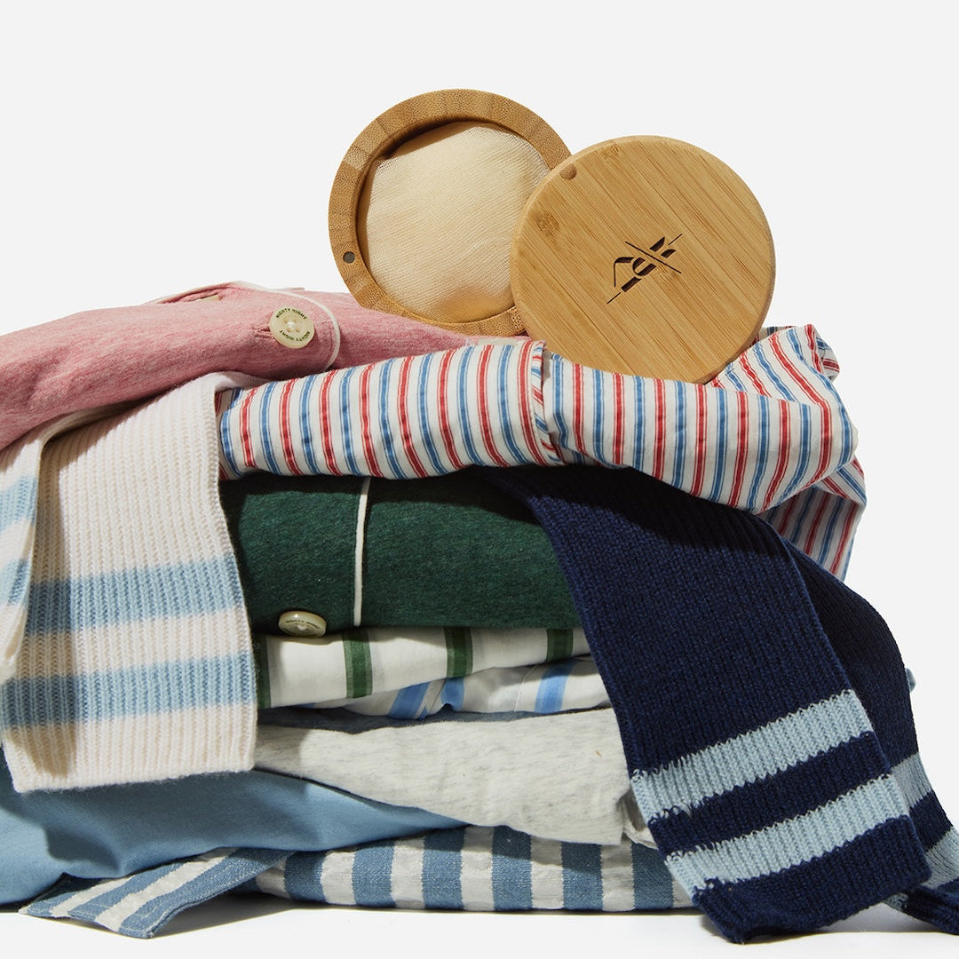 A styled flat-lay of folded knit clothing and accessories on a white background, including striped and solid knit garments in pink, red, blue, green, and navy, with two round light wood lids on top.
