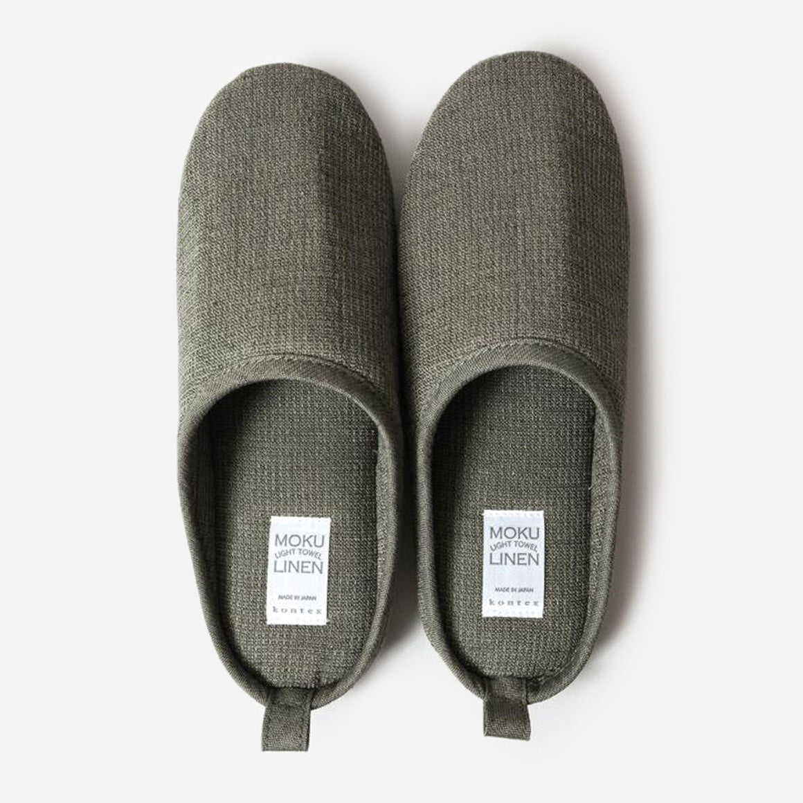 Top-down view of grey house slippers against white background. Moku Light Towel Linen written on white product tag on footbed.